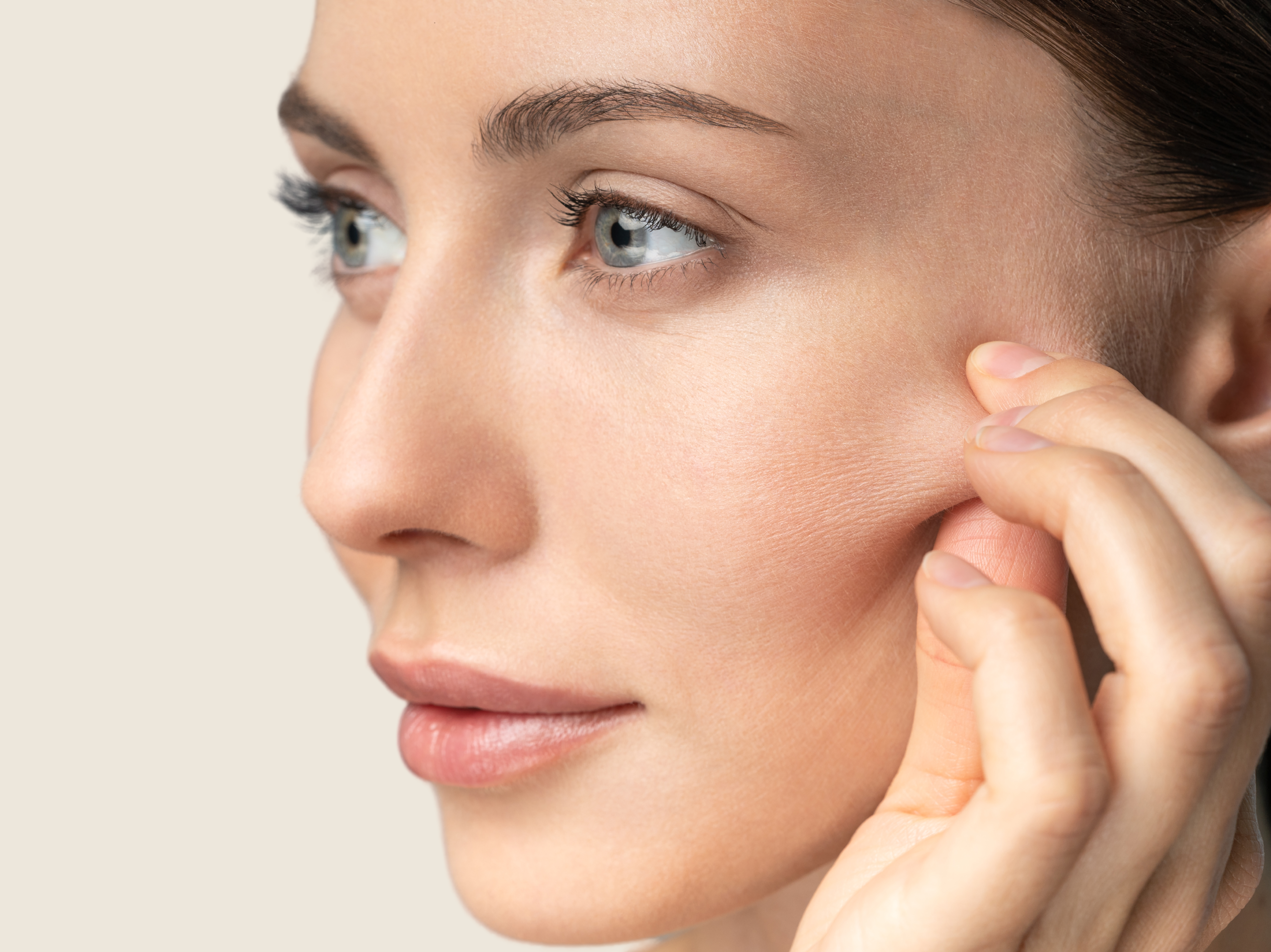 Blog non invasive Post Image Healthy beautiful Woman, side left face pinching her cheeks with beige background.